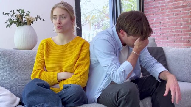 A young couple is experiencing relationship problems, sitting back to back on a couch, clearly upset after an argument. Their body language expresses sadness, conflict, and a lack of communication in 