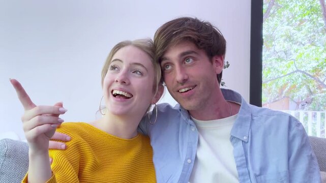 A happy young couple points and looks with excited expressions from their sofa. Sharing a joyful, heartwarming moment, they laugh together, embodying affection and connection in their home.