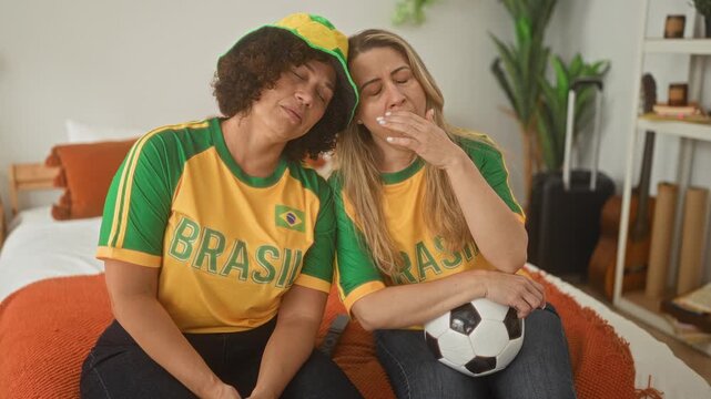 Woman in brasil jersey and hat holding a soccer ball with friend, cheering and jumping with raised arms on a bed in a building; celebration joy.