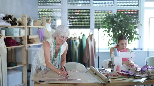 Adult woman seamstress sews on machine elderly woman assistant drawing pattern on paper in workshop