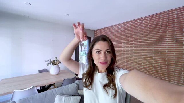 Happy young woman celebrating new home ownership, joyfully showing off her house keys in a modern apartment. Excited homeowner waving to the camera, sharing her successful real estate purchase.