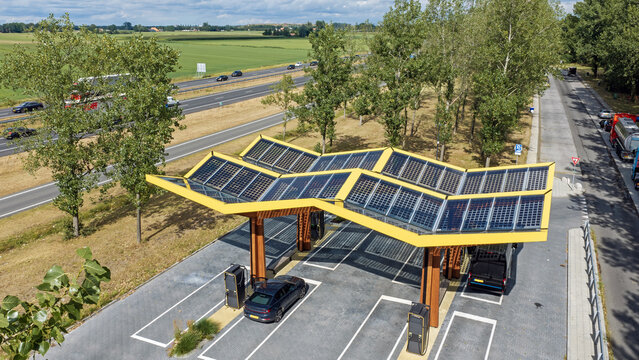 Aerial view of the Fastned charging station with its yellow solar canopy, electric vehicles parked at charging points, and a highway in the background in Haskerhorne, Friesland, Netherlands.