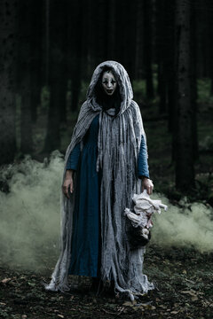 Spooky and scary Halloween themed image in a dark forest with smoke and an eerie woman with a mask. .