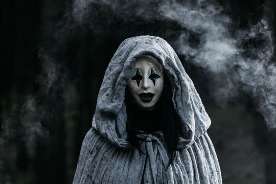 Spooky and scary Halloween themed image in a dark forest with smoke and an eerie woman with a mask. .