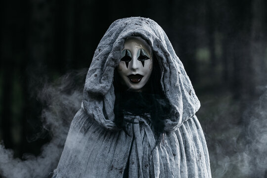 Spooky and scary Halloween themed image in a dark forest with smoke and an eerie woman with a mask. .