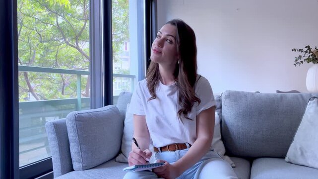 A young woman sits on a cozy sofa, jotting down thoughts in a notebook. She looks contemplative and inspired, enjoying a moment of quiet reflection and planning in her modern, sunlit apartment.