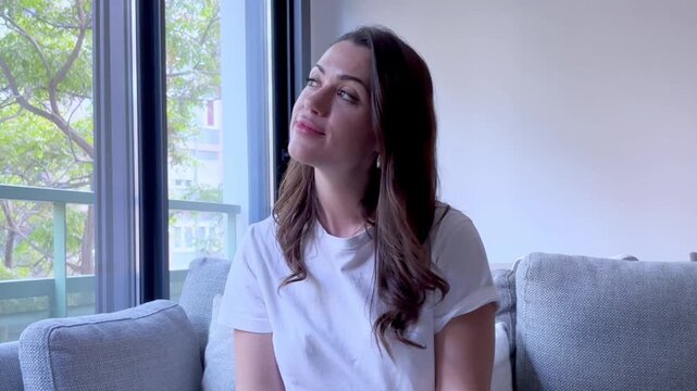 A serene young woman enjoys peaceful relaxation at home, sitting on a comfortable sofa. With closed eyes, she feels calm and content, appreciating the quiet ambiance of her modern living space.