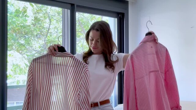 A young woman decides what to wear, holding up two striped shirts in her bright, modern bedroom. She thoughtfully compares the outfits, making a fashion choice for her day.