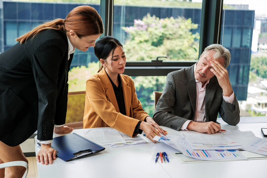 Three colleagues in an office review financial reports and charts; one looks stressed while others discuss solutions around a conference table by a window