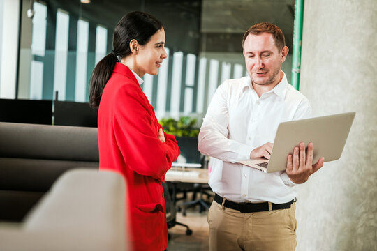 Two professionals in a modern office discuss a project while reviewing a laptop, suggesting collaboration, strategy, and teamwork.