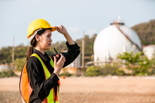 A worker wearing a hard hat and safety vest uses a two-way radio at an industrial site, with a spherical storage tank in the background.