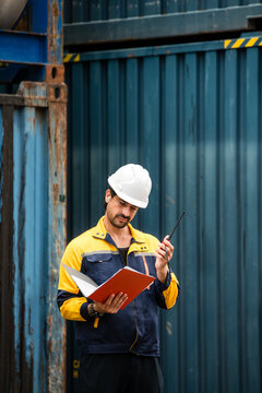 Worker in hardhat checks documents and talks on a radio beside stacked shipping containers at a freight terminal.