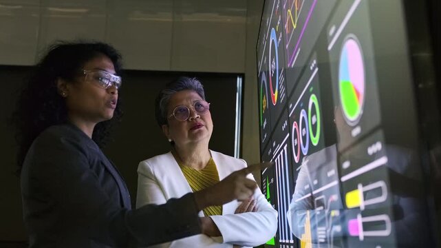 Two professionals review interactive data dashboards on a large touchscreen, analyzing charts and metrics during a business meeting