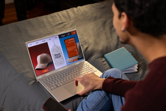 African American man browsing Black Friday on bed using silver laptop and holding phone, copy space