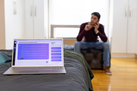 African American man sitting on ottoman using phone in bedroom with laptop showing marketing plan