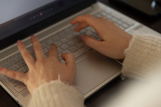 Woman typing on silver laptop on dark wood desk wearing fuzzy sweater, showing pink nails