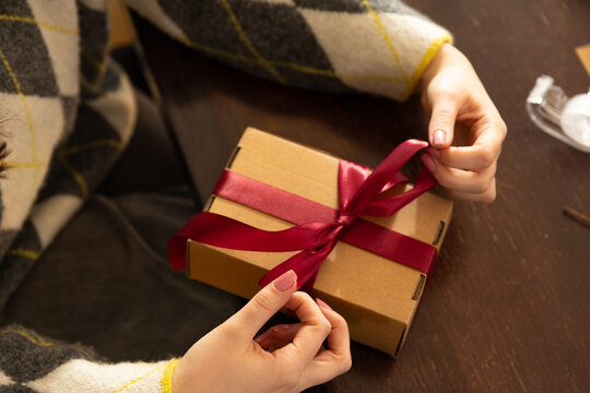 Adult female in argyle sweater tying burgundy ribbon on box at wooden table with tape dispenser
