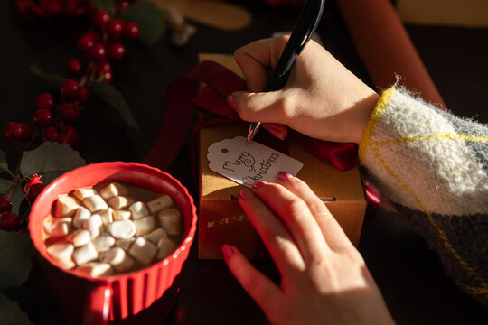 Adult woman wearing striped sweater writing tag with pen holding ribbon gift box at home table