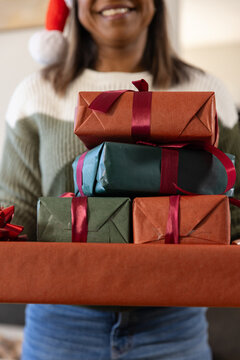 Mature African American woman holding wrapped presents with red bow at home wearing Santa hat