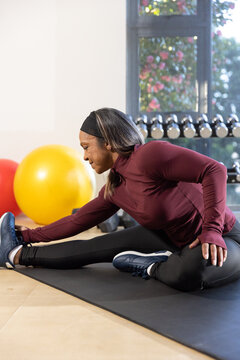 Mature woman stretching on black mat in fitness room wearing maroon top with dumbbells nearby