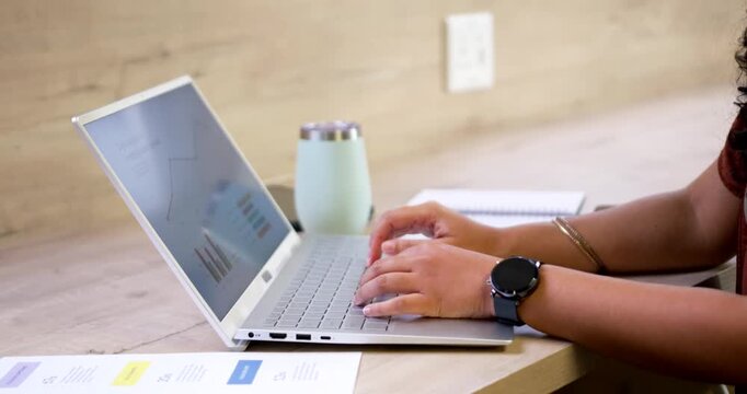 African American woman noticing printed charts on silver laptop screen, typing at desk, copy space