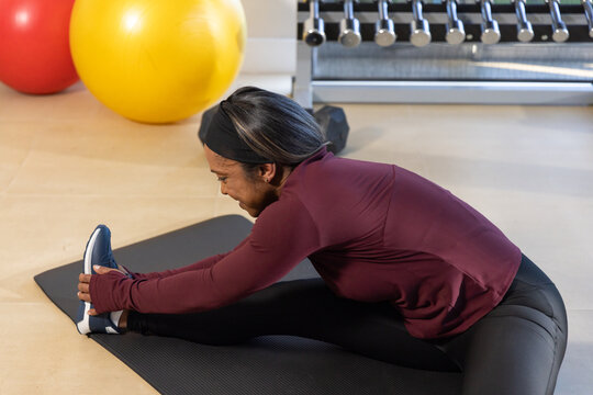 African American woman performing forward stretch on mat in gym near balls wearing maroon top