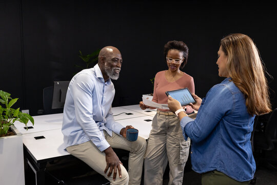 Diverse coworkers meeting at white desks in office attire, holding tablet, papers and blue mug
