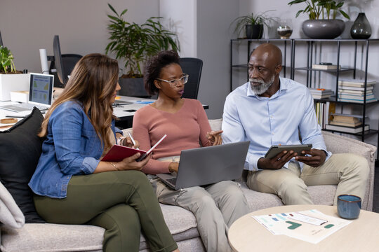 Diverse coworkers in work attire discussing project on office sofa with laptop tablet red notebook