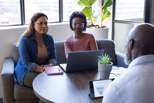 Diverse coworkers discussing project in office lounge around round table with laptop, tablet