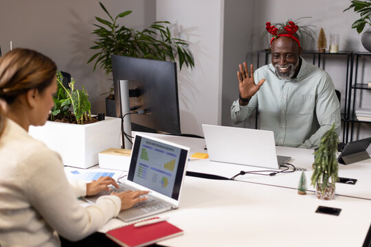 Diverse coworkers waving, typing at open desk with laptops, plants and red antlers, copy space