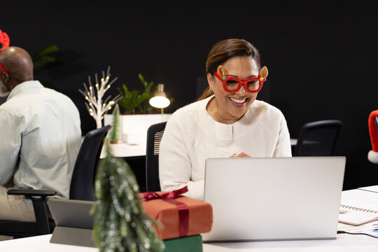 African American coworkers decorating desk with gifts, woman wearing reindeer glasses typing laptop