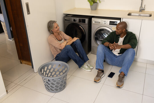 Couple sitting on tiled laundry room floor in jeans, talking beside front-loading washer and basket