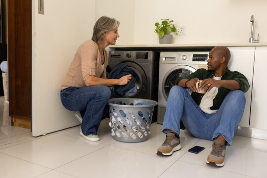 Couple loading clothes into washer and dryer in laundry room, gray basket, mug, smartphone nearby