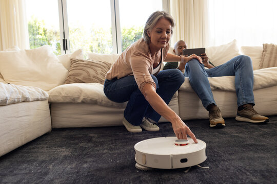Couple senior woman crouching, pressing robot vacuum on dark rug at home while partner using tablet