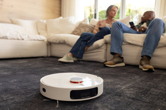 Senior woman and African American man holding phone and talking while robot vacuum cleaning carpet