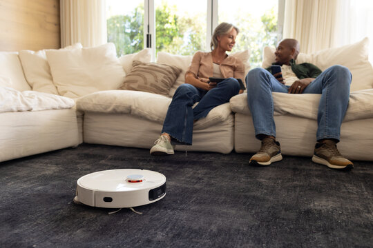 Diverse couple sitting on sectional sofa at home using phones while robot vacuum cleaning rug