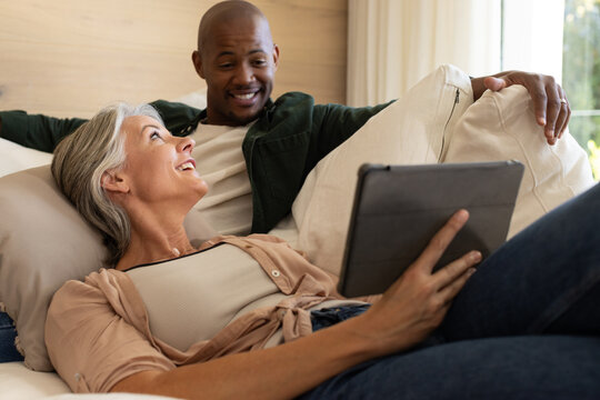 Diverse couple with African American man and senior woman lounging on sofa holding tablet