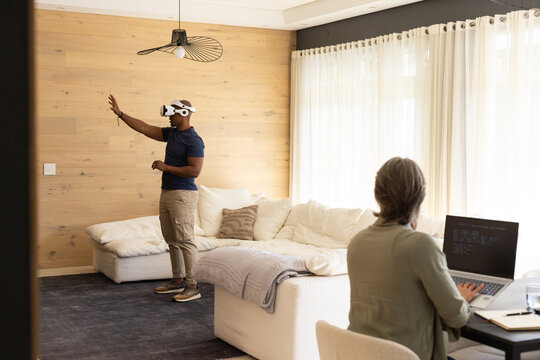 Couple man wearing VR headset reaching in living room while woman coding on laptop, copy space