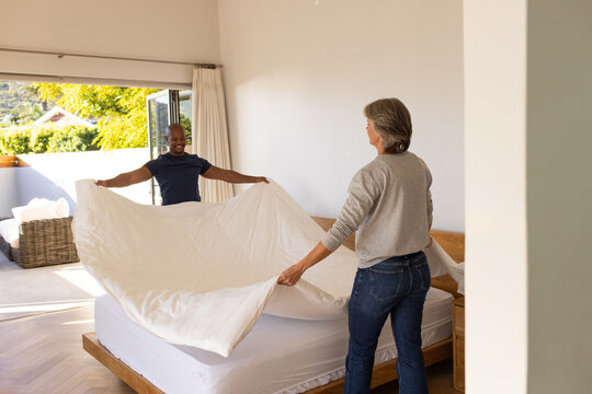 Senior woman and African American man spreading white duvet on platform bed by open patio door