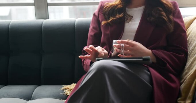 Adult female in blazer holding glass cup, tapping tablet resting on lap in lounge for work