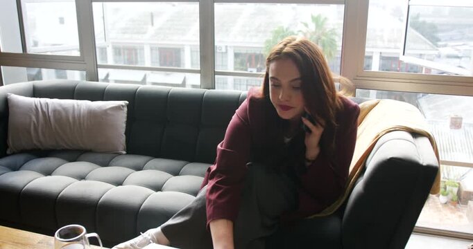 Woman in maroon blazer talking on phone, reaching laptop and reading chart for work on sofa