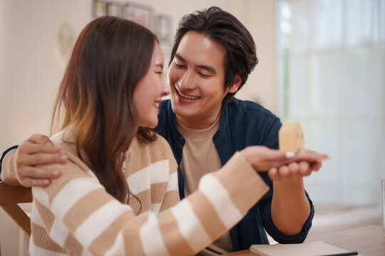 Happy couple smiling together while holding a small house model at home, representing home ownership, property purchase, mortgage planning, real estate investment and a shared future.