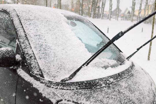 Close-up of a snow-covered automobile front window and lifted wiper arm after winter blizzard. Frozen vehicle on outdoor parking lot, cold weather and snowfall concept.