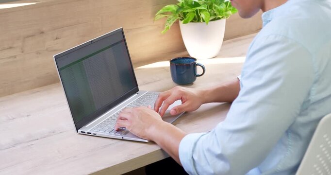 Asian man typing spreadsheet on laptop at desk, pausing work, sipping from blue mug, copy space