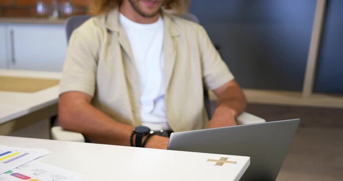 Camera pulling back revealing man adjusting posture, leaning forward, typing on laptop at office
