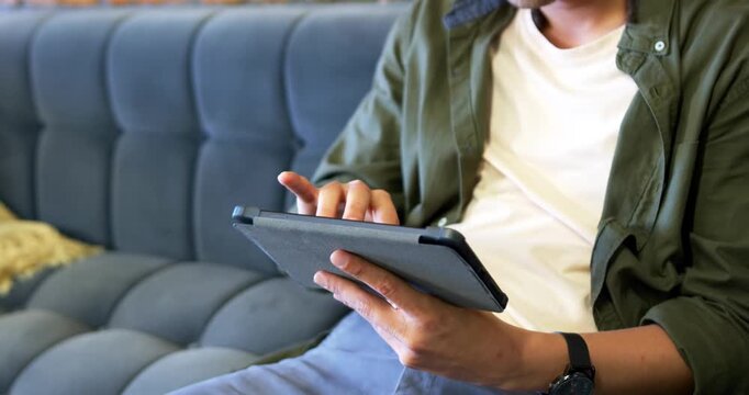 African American man starting by resting tablet on lap, sitting on tufted blue sofa, tapping screen