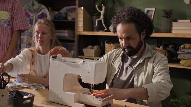 Middle aged man using electric sewing machine to stitch fabric in workshop while young adult Caucasian woman works in background. Clip is perfect for promoting tailoring classes