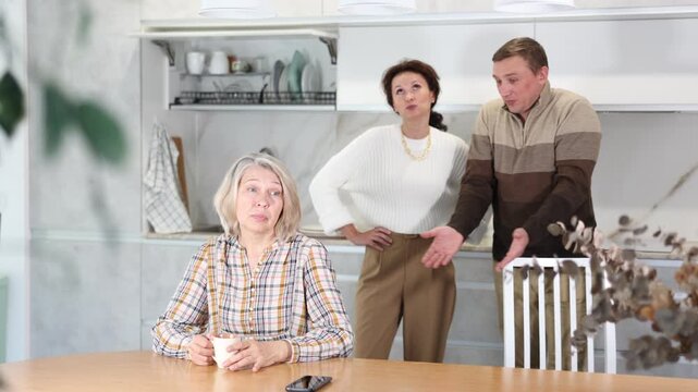 Upset old woman sitting at table with her back to angry middle-aged man and woman quarreling to her in the kitchen. High quality 4k footage