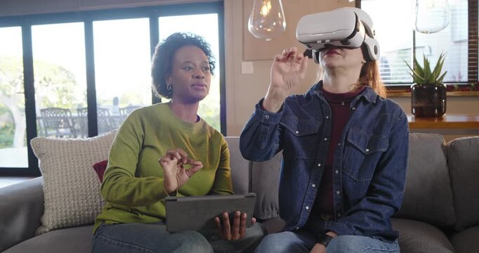 Diverse female friends tapping tablet, guiding VR user reaching and selecting virtual items on sofa