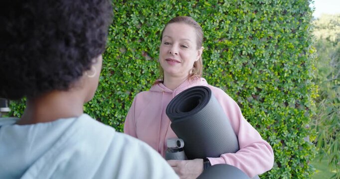 Diverse female friends meeting for yoga, pink-hoodie woman coming with two mats and bottle laughing